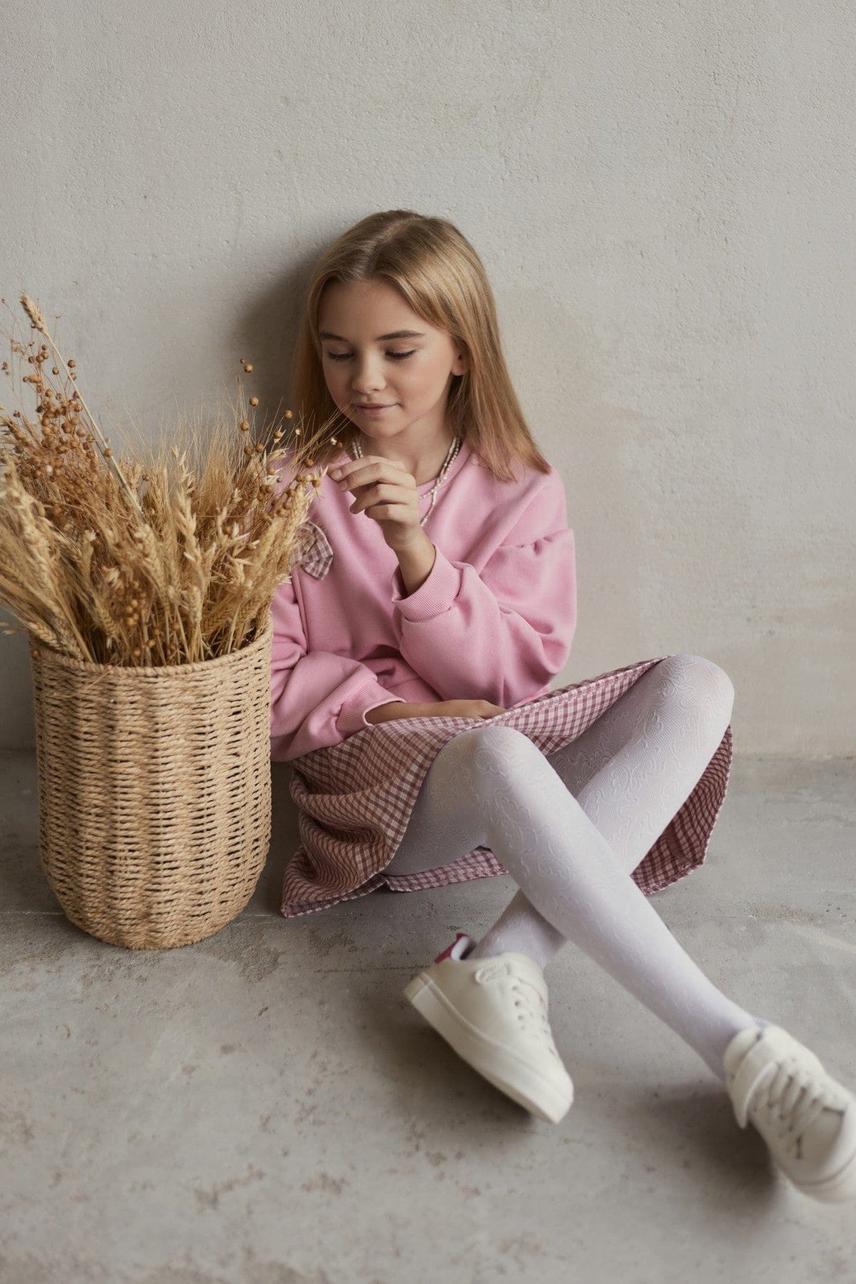 A young girl with blonde hair sits on the floor wearing a pink sweatshirt, a checkered skirt, white patterned tights, and white sneakers next to a tall wicker basket filled with dried wheat.