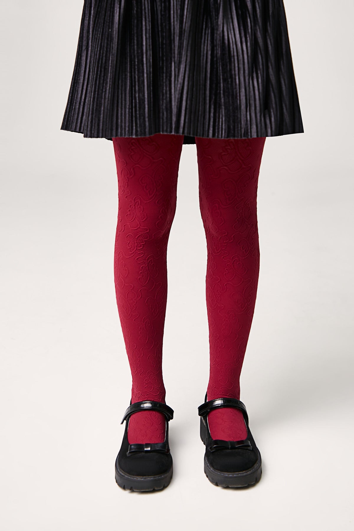 A pair of red patterned tights worn with a black pleated skirt and black Mary Jane shoes featuring a bow detail.