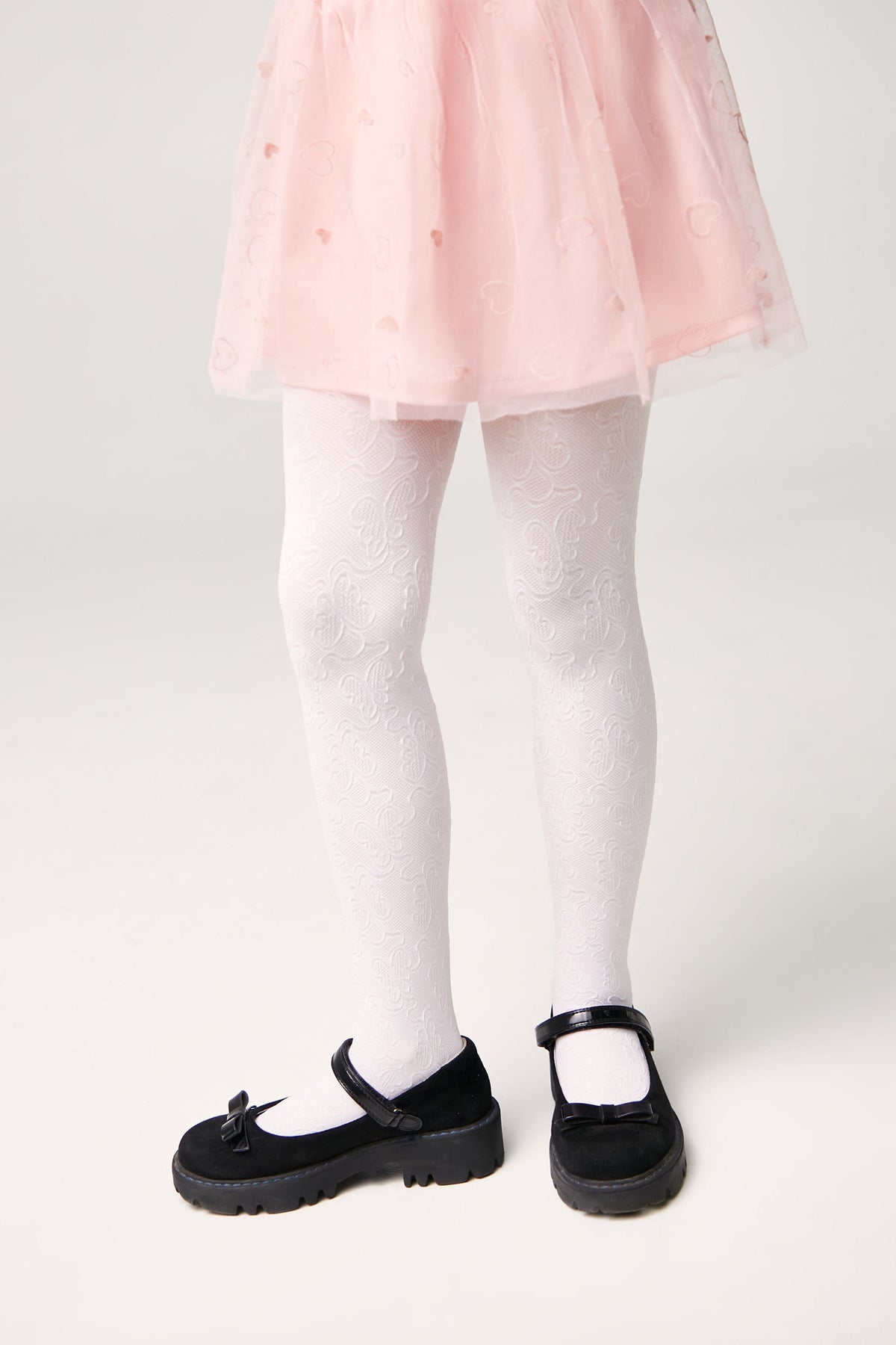 A child wears white patterned tights with a light pink tutu skirt and black Mary Jane shoes.