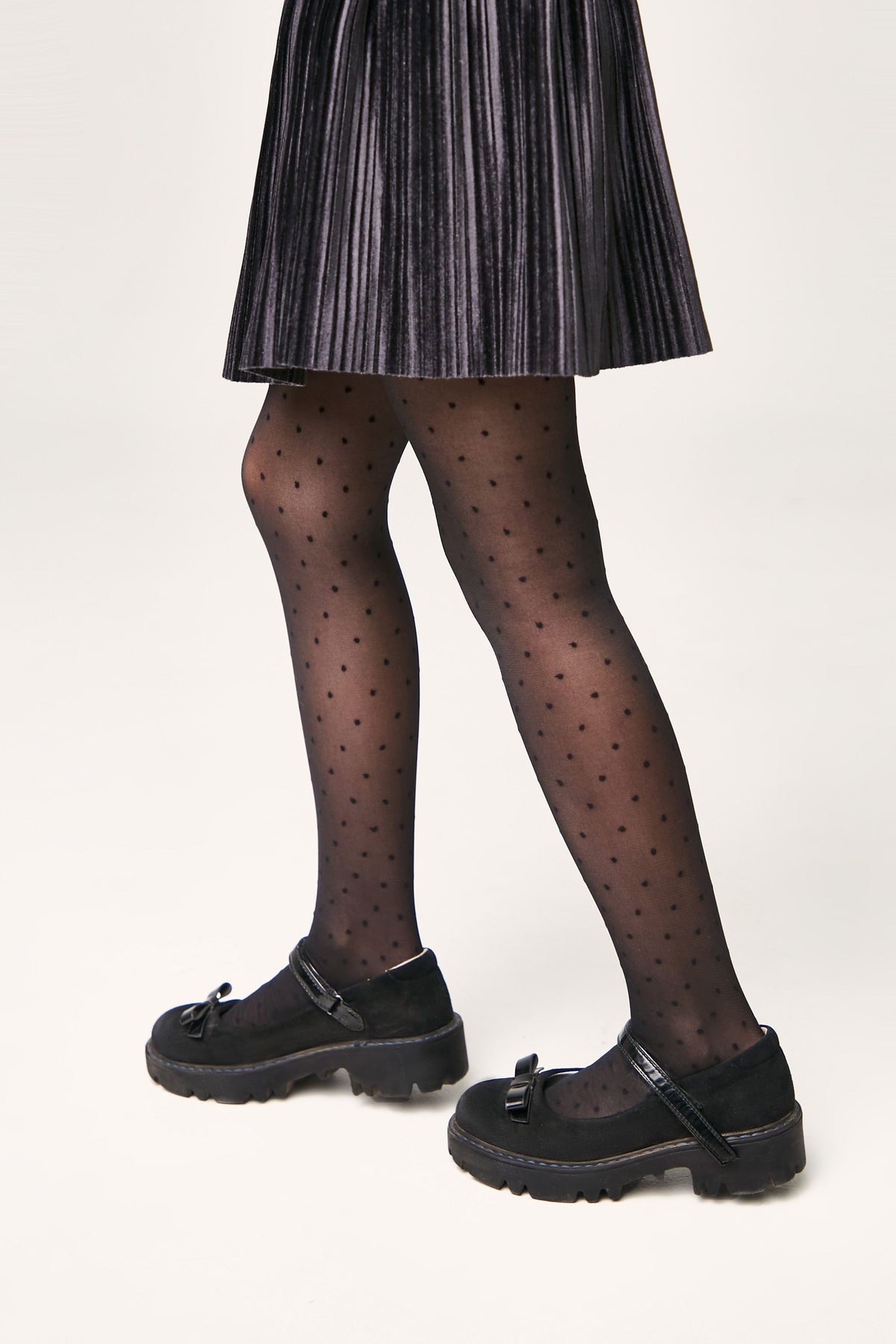 A person wears a pleated black skirt, sheer black tights with a small polka dot pattern, and black platform Mary Jane shoes with buckled straps and ribbon accents.