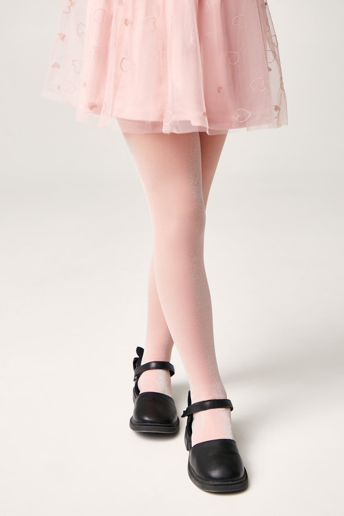 A child wears light pink, glittery tights paired with a pink heart-patterned tulle skirt and black Mary Jane-style shoes.