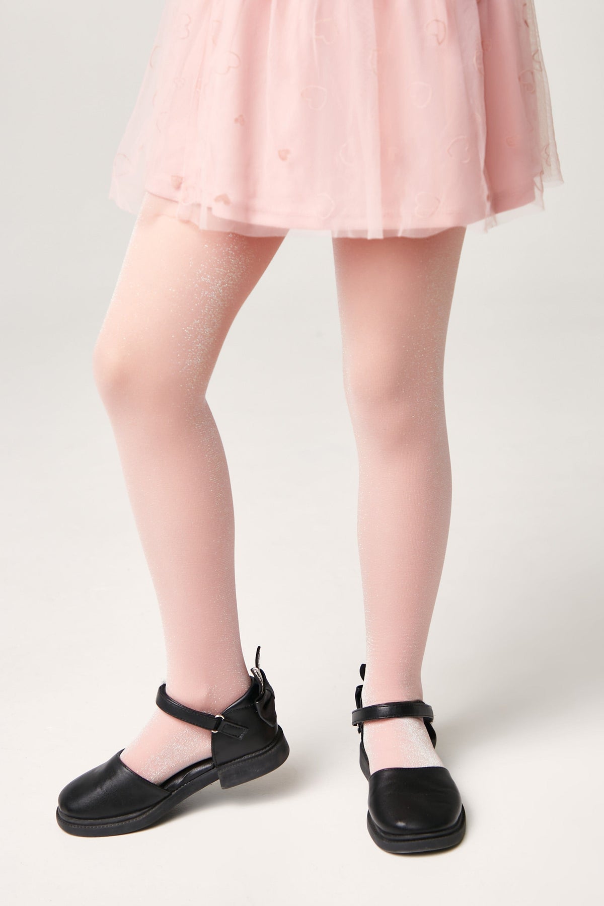A child wears pale pink glittery tights paired with black mary jane shoes under a light pink skirt.