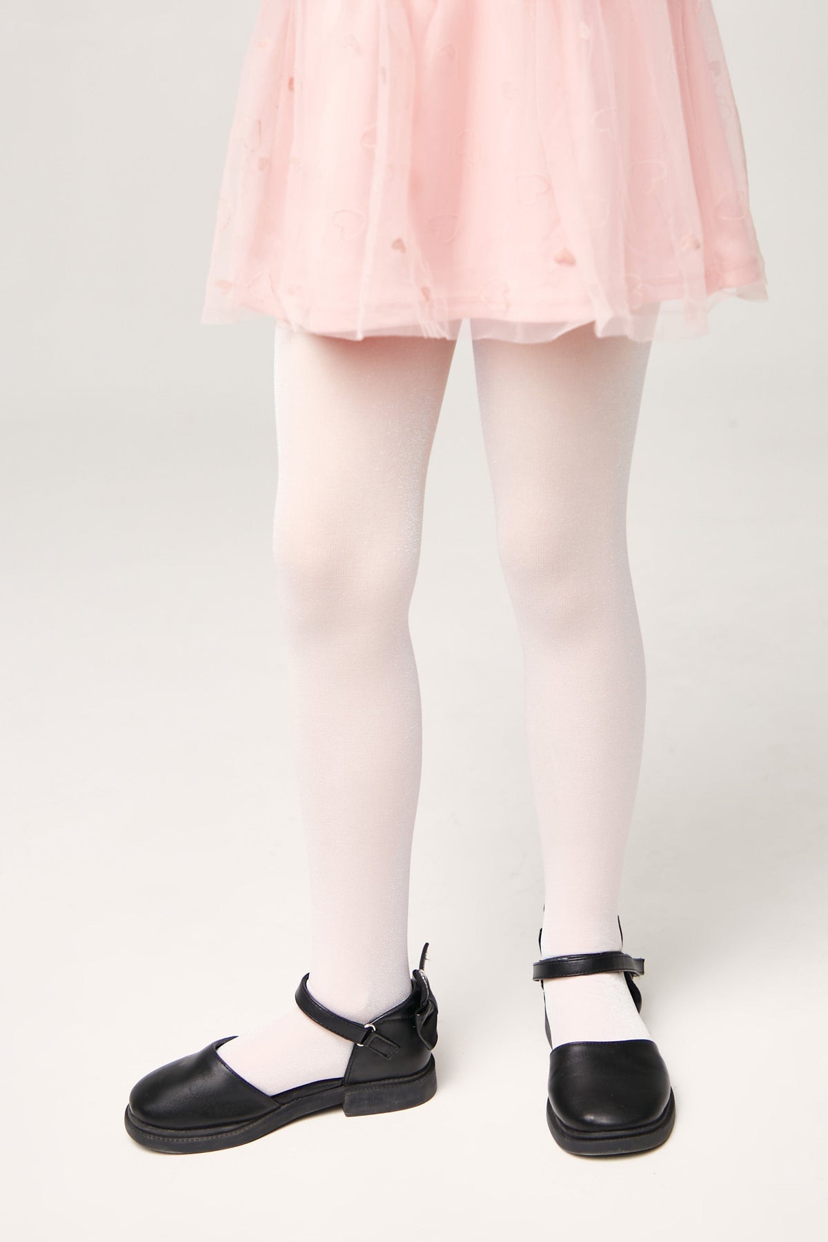 A pair of white tights worn with a light pink tulle skirt and black Mary Jane shoes on a plain light background.