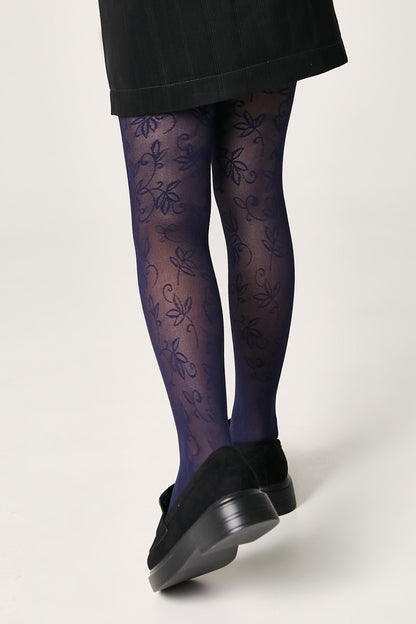 A person wears sheer, navy blue patterned tights with a delicate floral design paired with black loafers and a dark skirt against a plain background.
