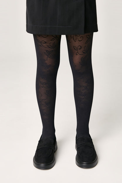 Black patterned tights featuring an intricate floral vine design worn with a black skirt and matching black loafers against a plain white background.