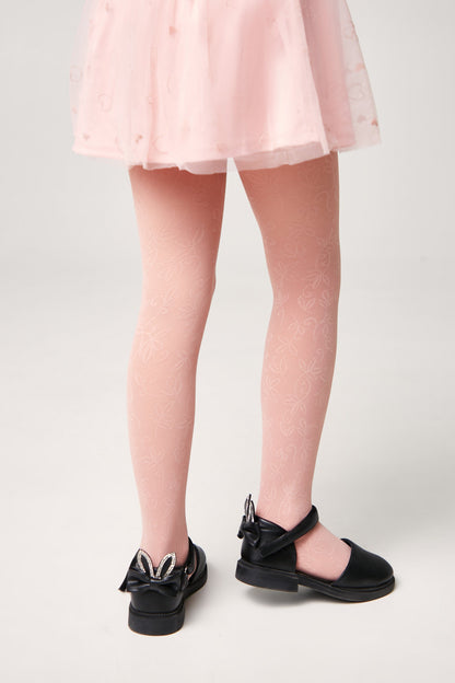 A child wears pale pink patterned tights and black mary jane shoes with decorative bunny ear bows on the heels, paired with a light pink tulle skirt.