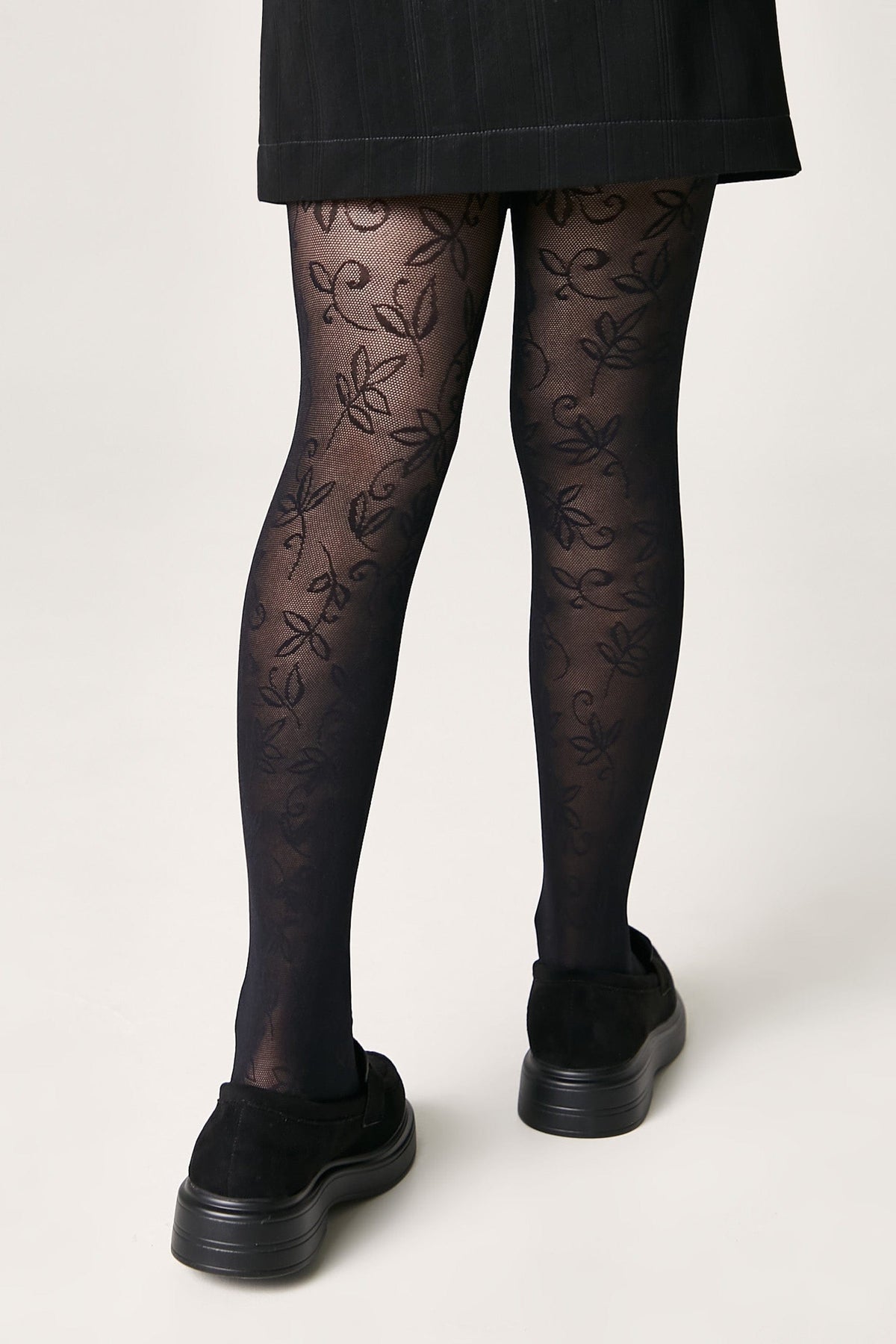 A pair of black patterned tights with a delicate leaf design worn under a black skirt and paired with black platform slip-on shoes.