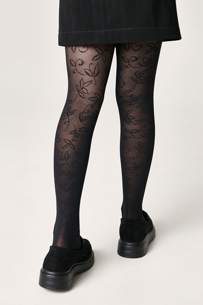 A pair of black patterned tights with a delicate leaf design worn under a black skirt and paired with black platform slip-on shoes.
