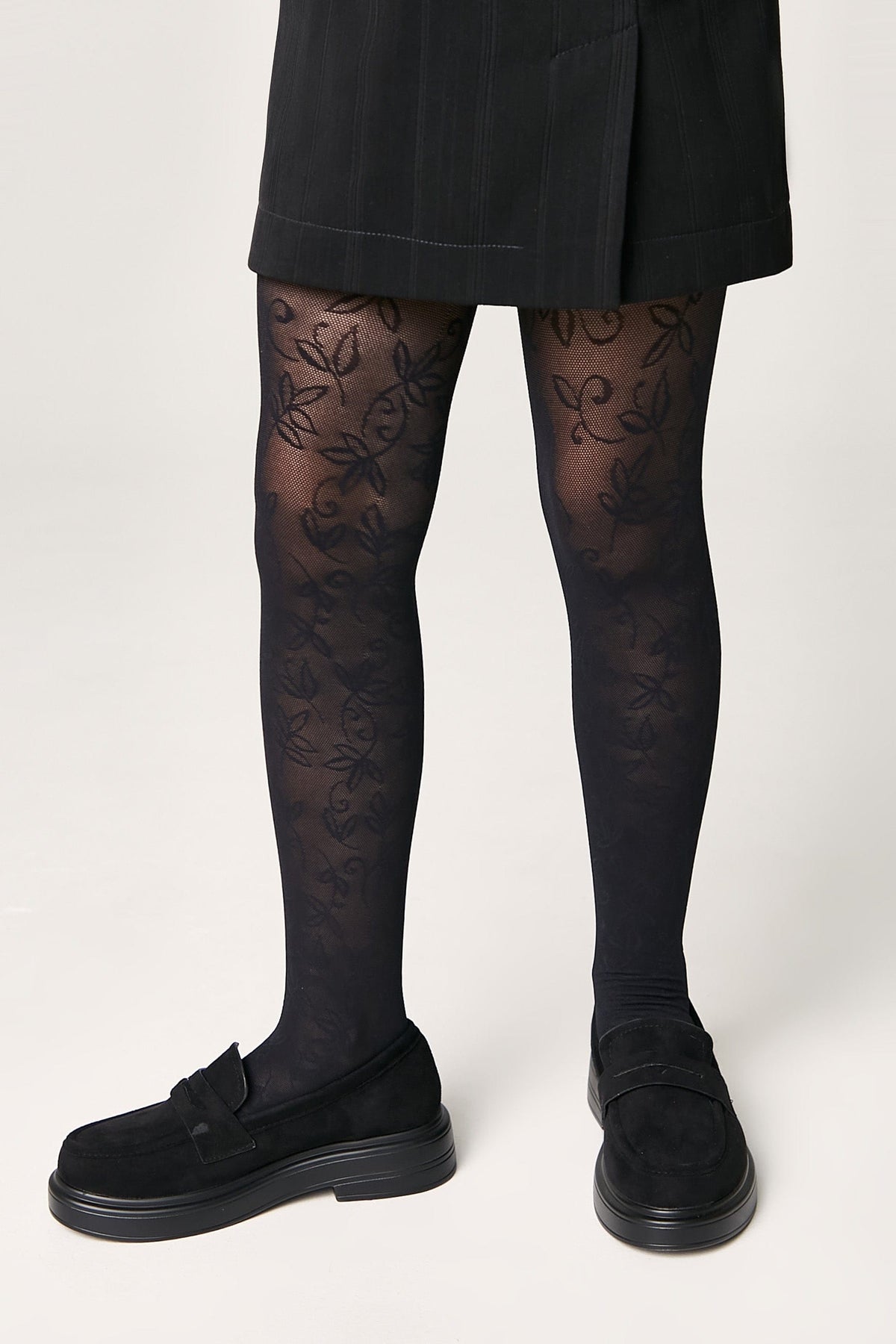 Black patterned tights featuring a delicate floral vine design worn with a black skirt and black suede-like loafers.