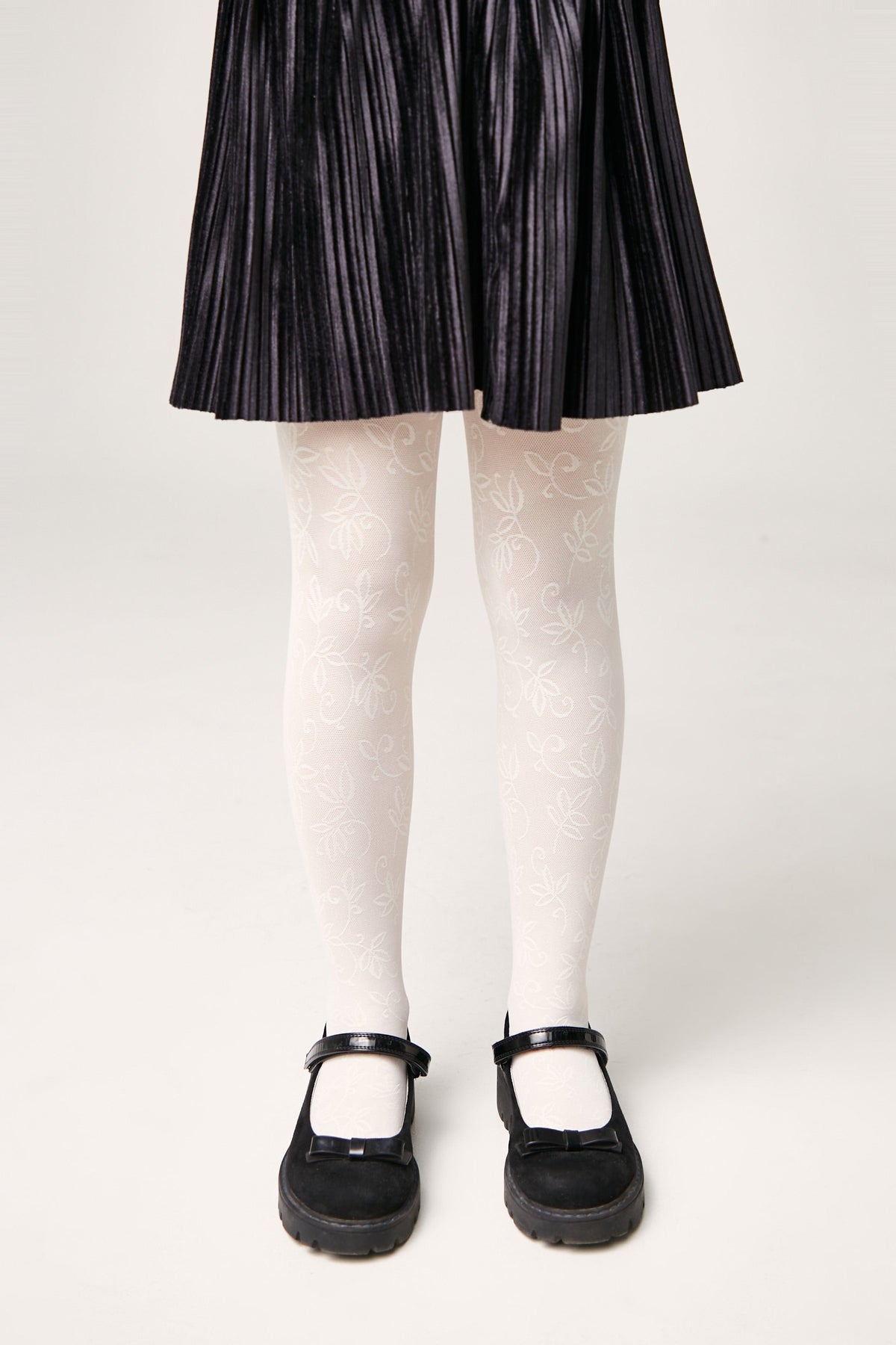 A child wears white tights with a subtle leaf pattern paired with a black pleated skirt and black Mary Jane shoes.