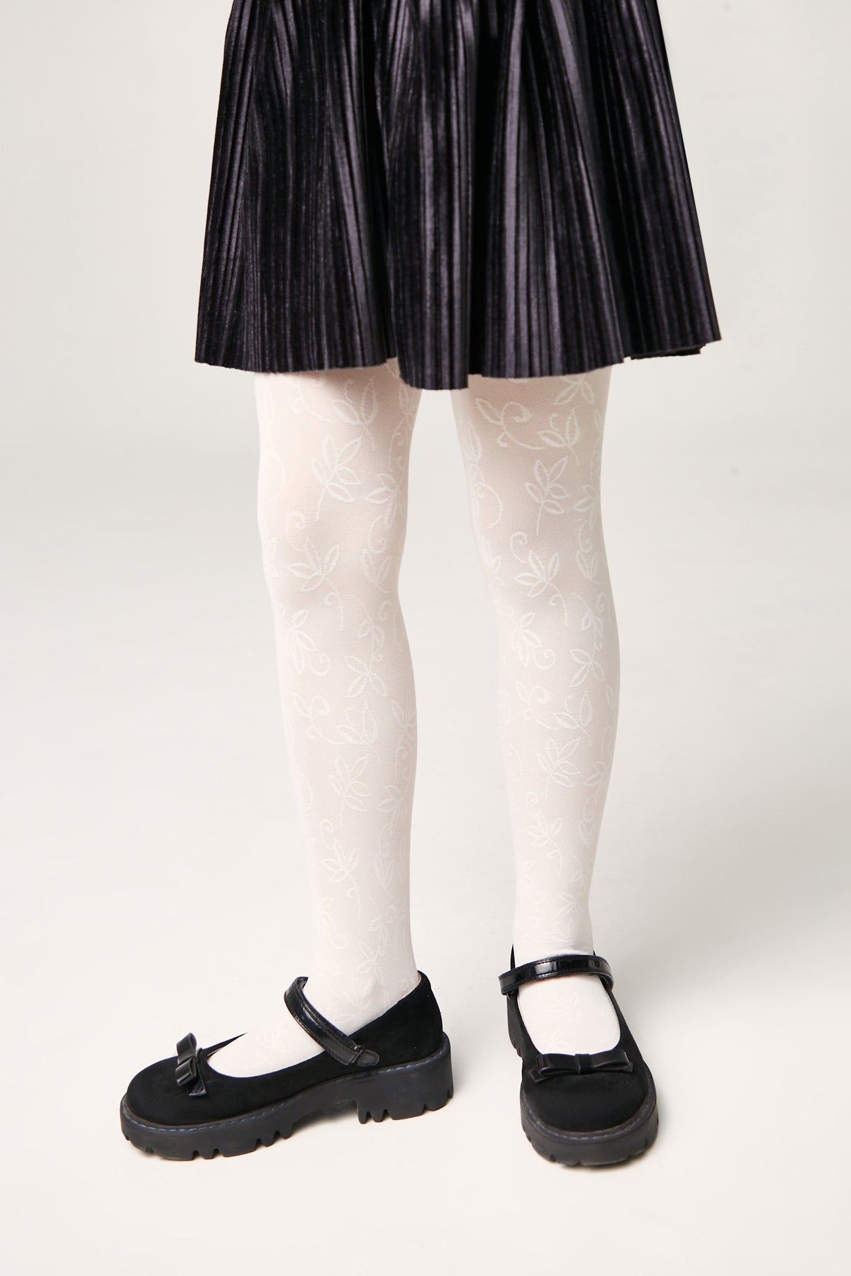 White tights with a delicate leaf pattern worn with a black pleated skirt and black mary jane shoes with bow details.