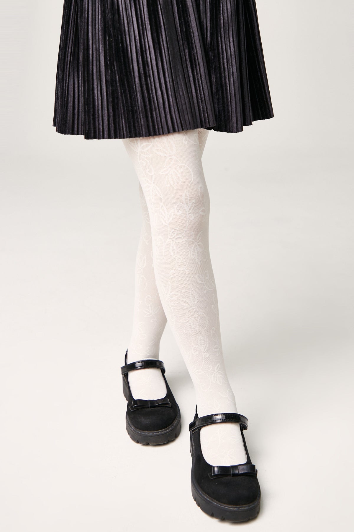 White patterned tights with a delicate leaf design worn with a black pleated skirt and black Mary Jane shoes featuring small bows.