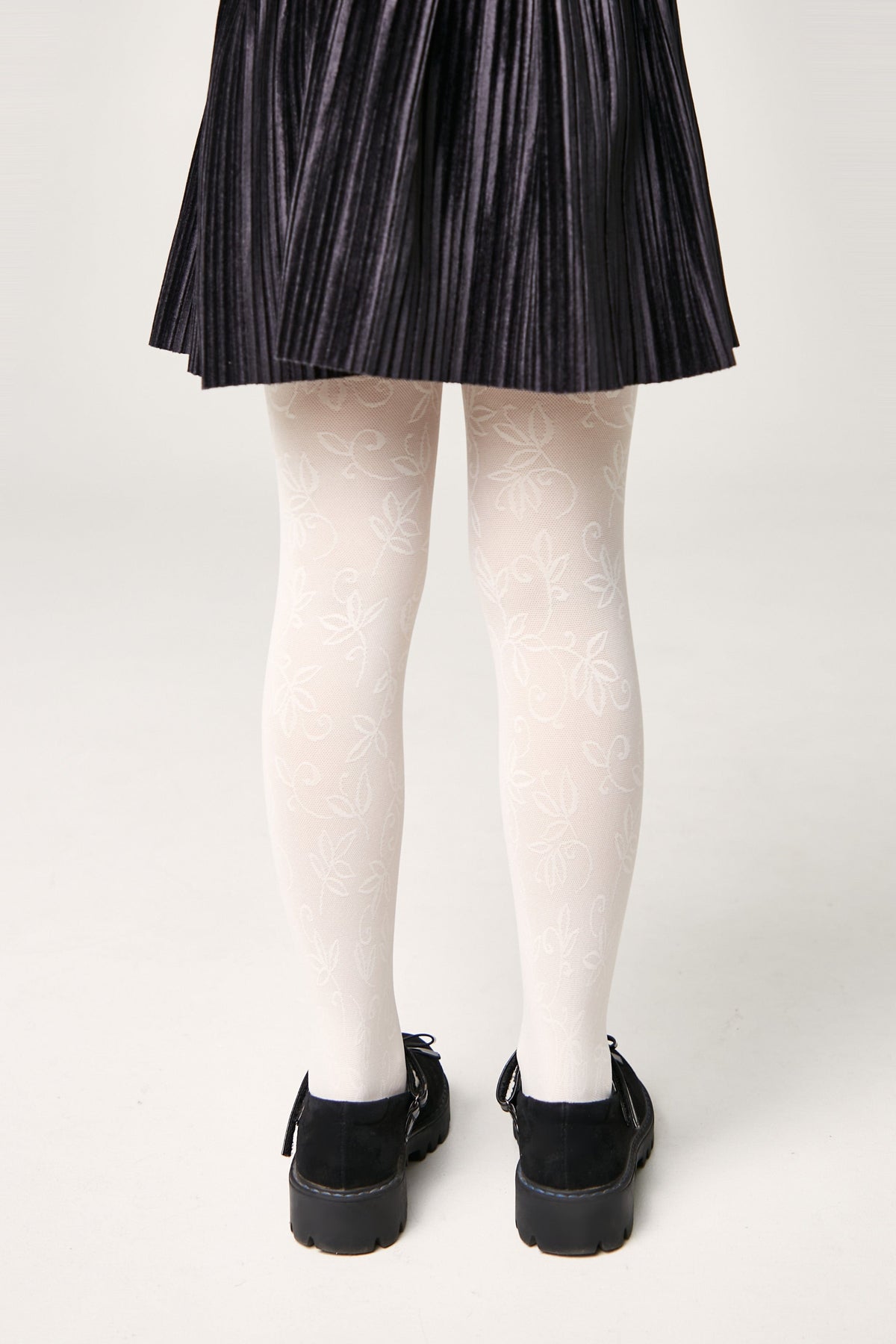 A person wears white patterned tights featuring a repeating floral leaf design, paired with a black pleated skirt and black buckled shoes.
