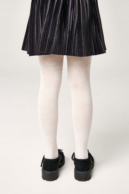 A person wears white patterned tights featuring a repeating floral leaf design, paired with a black pleated skirt and black buckled shoes.