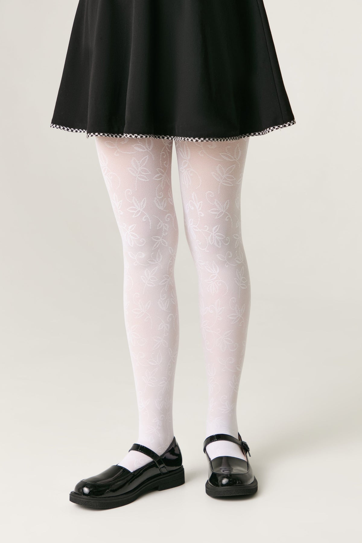 A pair of white tights featuring a delicate, subtle leafy vine pattern, worn with a black skirt and glossy black Mary Jane shoes against a plain grey background.