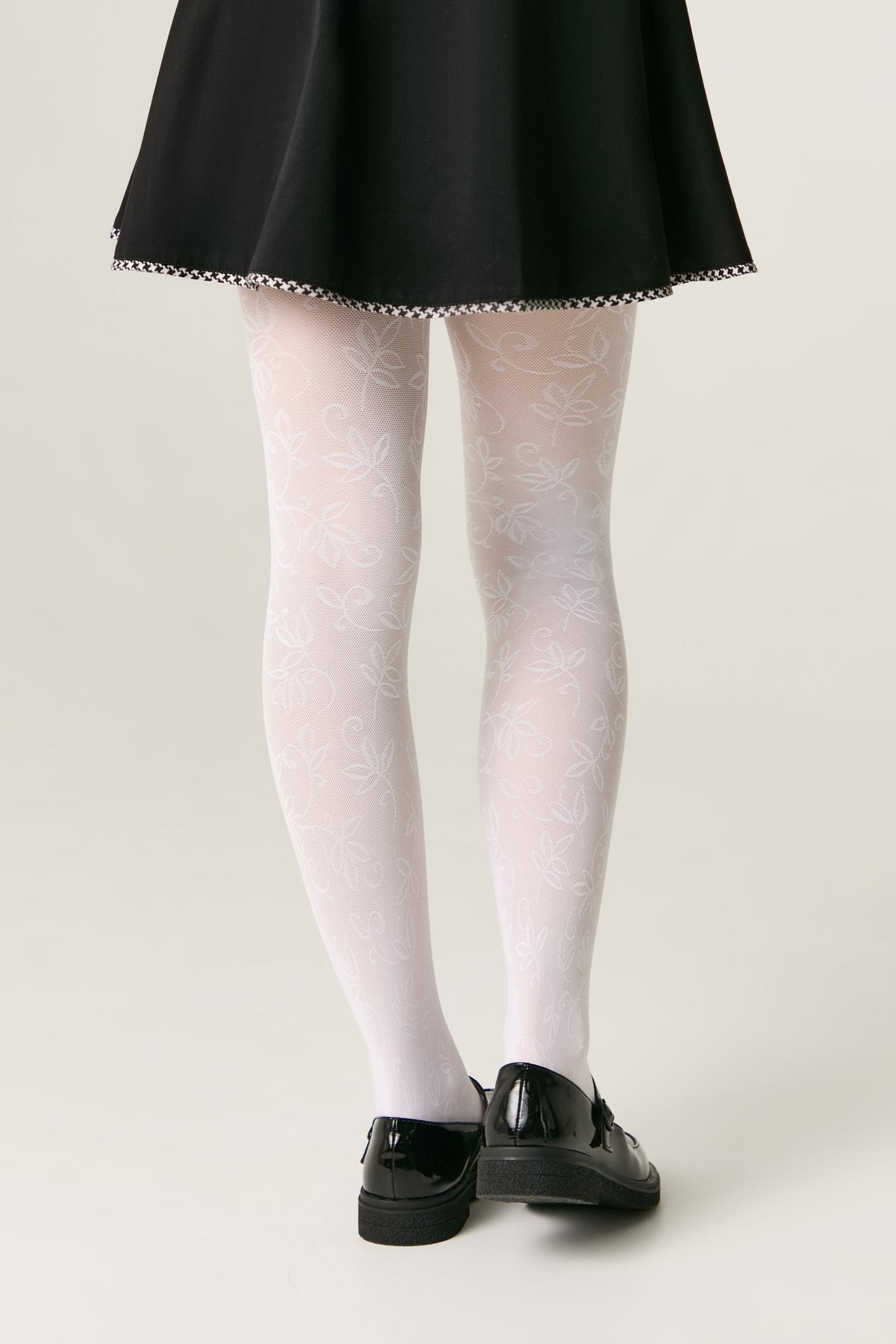 White patterned tights with a delicate leaf and vine motif worn under a black skirt with patent black flat shoes against a plain background.