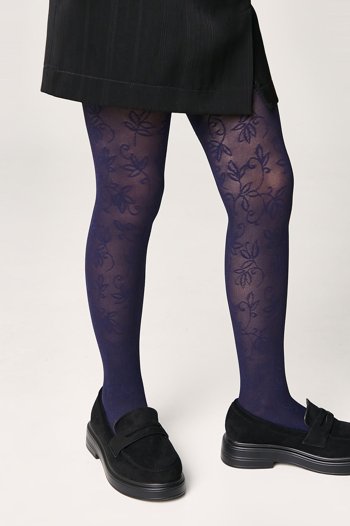 A pair of deep blue patterned tights featuring a delicate floral vine design, worn under a black skirt with black platform loafers.