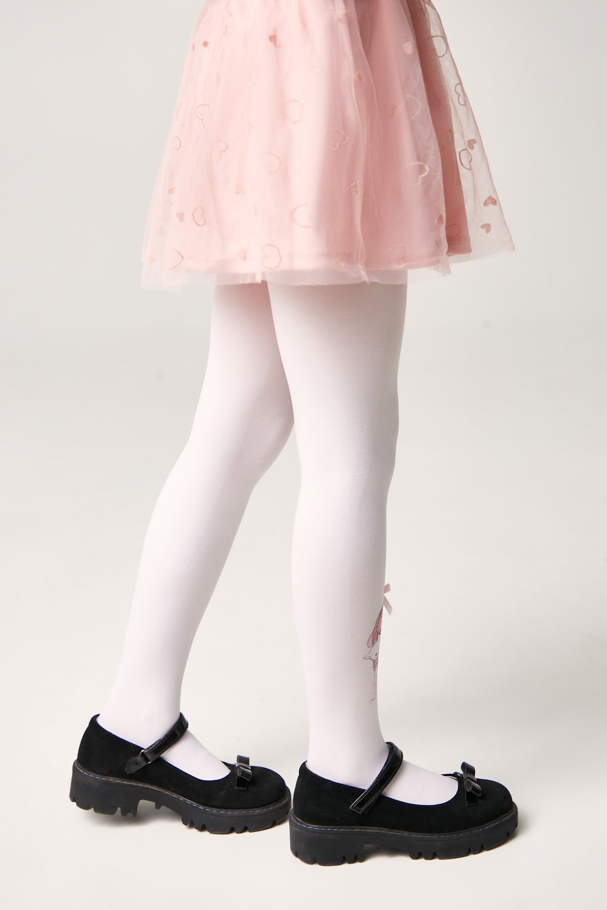 A pair of white tights with a small pink heart detail on the side, worn with a pink heart-patterned skirt and black mary jane shoes.