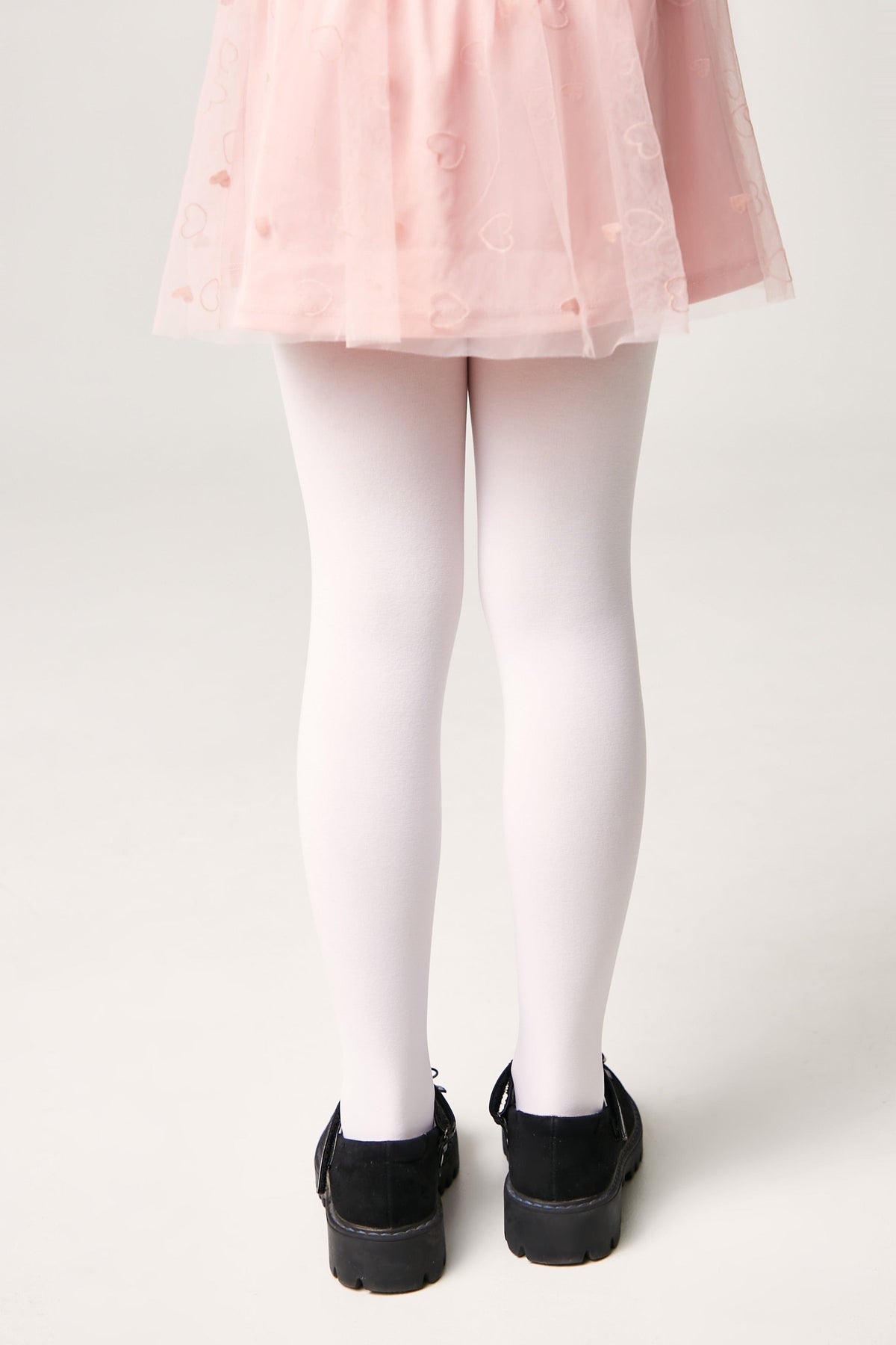 A pair of opaque white tights worn with a light pink tulle skirt featuring a heart pattern and black buckled shoes.