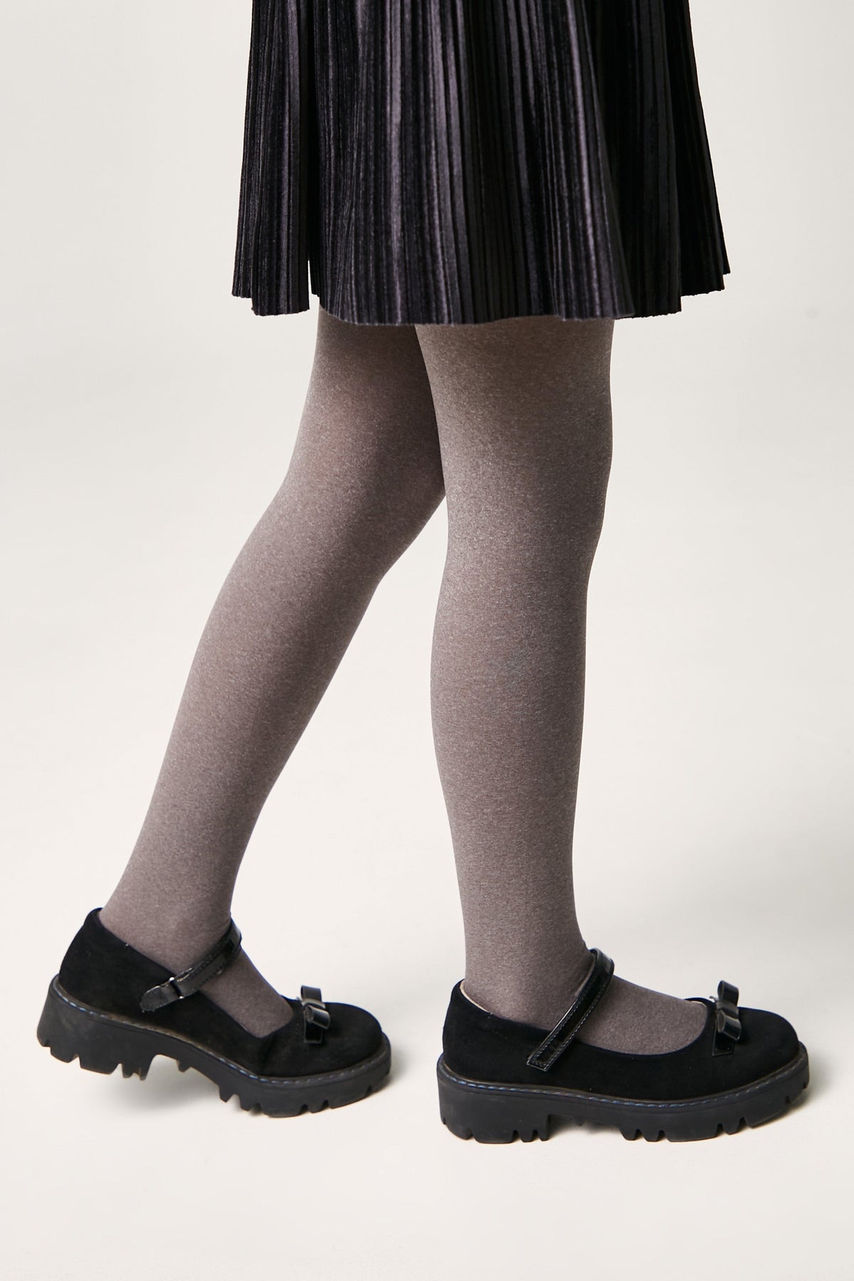 A pair of black suede Mary Jane shoes with thick lug soles and a small bow detail on the front, worn with grey tights and a black pleated skirt.