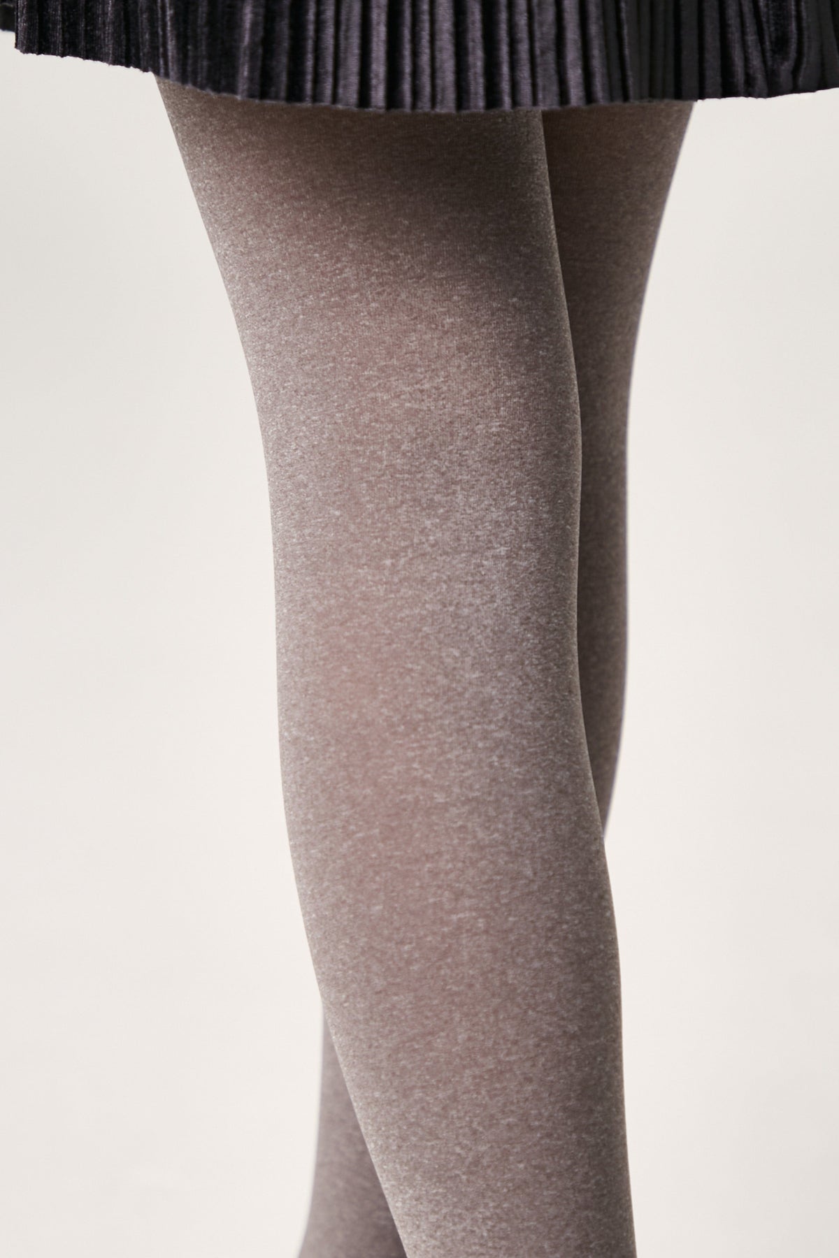 A close-up view of legs wearing opaque, heathered grey tights beneath a dark pleated skirt against a plain, light-colored background.