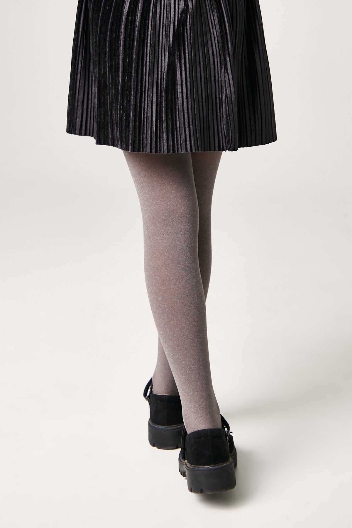 A pair of opaque grey tights worn with a black pleated skirt and black buckled flat shoes against a plain white background.