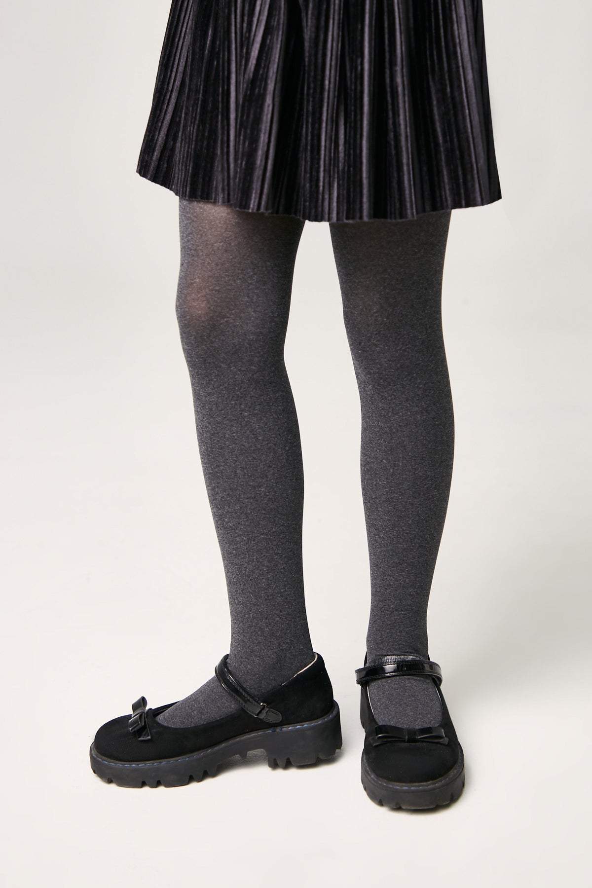A person wears a dark grey pleated skirt with matching grey tights and black flat shoes featuring a decorative bow and an adjustable strap.