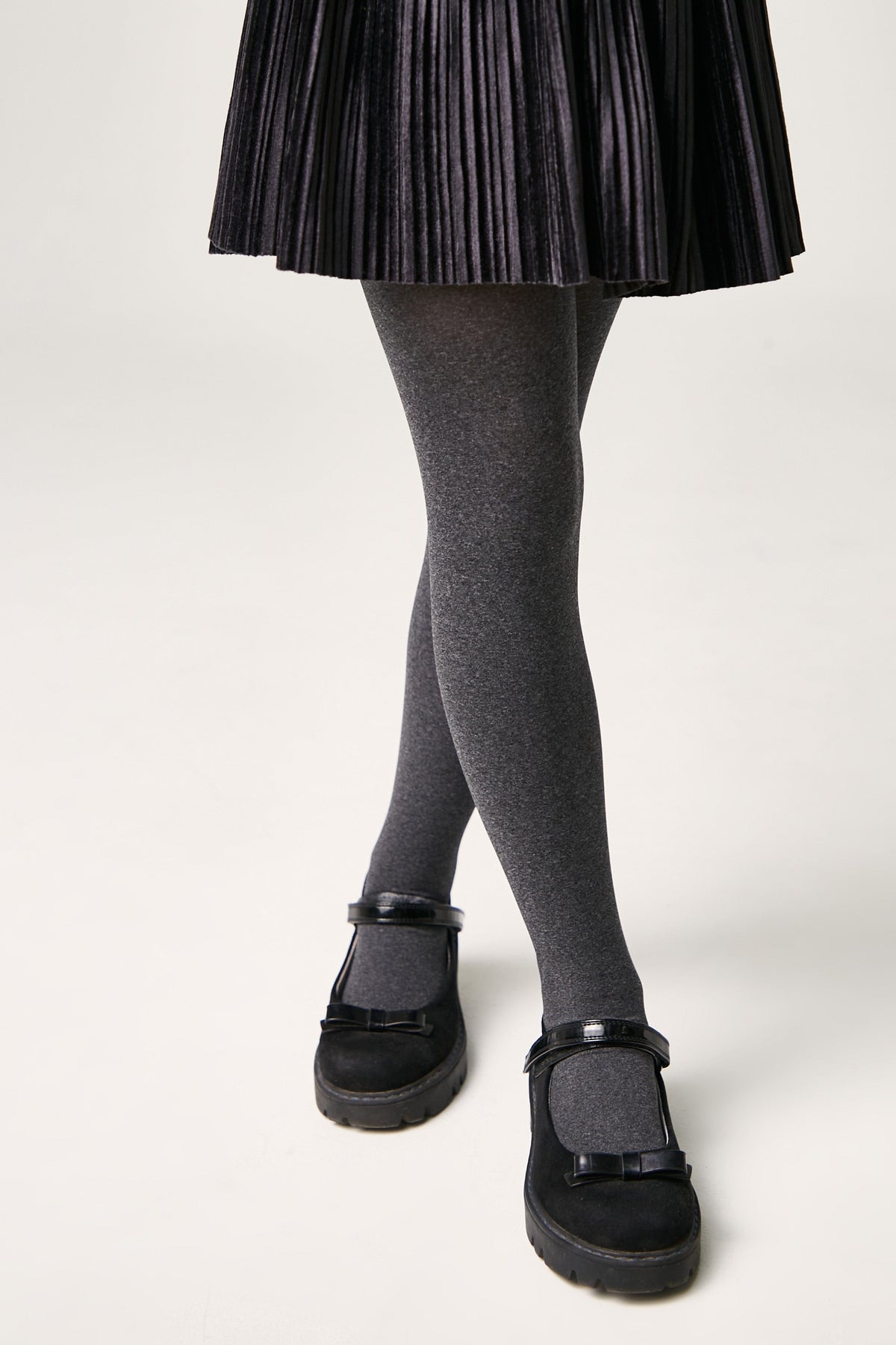 A person wearing a black pleated skirt, grey tights, and black Mary Jane shoes with bow details against a plain white background.