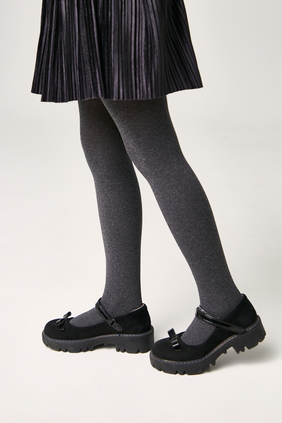 Black Mary Jane shoes with thick lug soles and small decorative bows on the straps, worn with grey tights and a pleated black skirt.