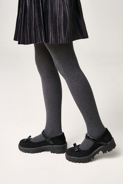 Black Mary Jane shoes with thick lug soles and small decorative bows on the straps, worn with grey tights and a pleated black skirt.
