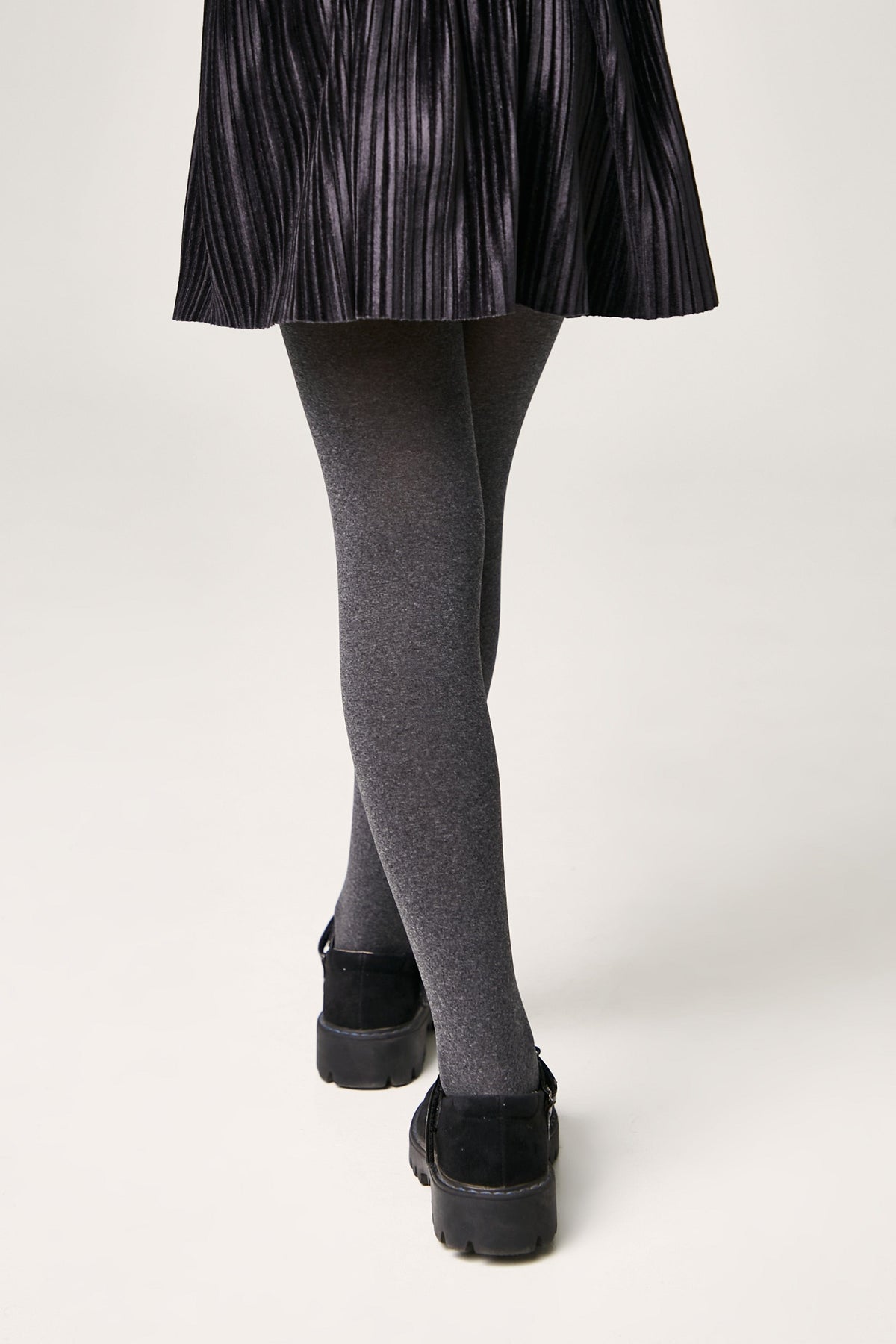A person wears a black pleated skirt with opaque grey tights and black flat shoes against a light grey background.