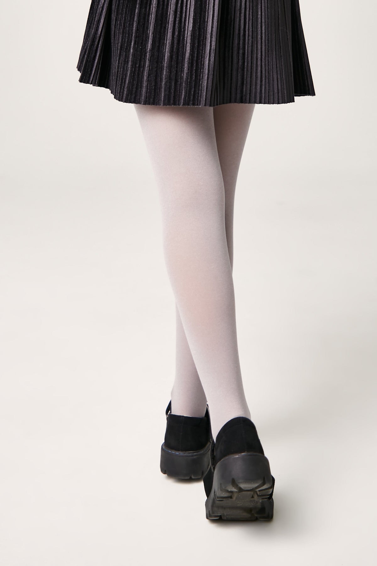 A person wears a black pleated skirt, light grey opaque tights, and black platform shoes with lug soles.