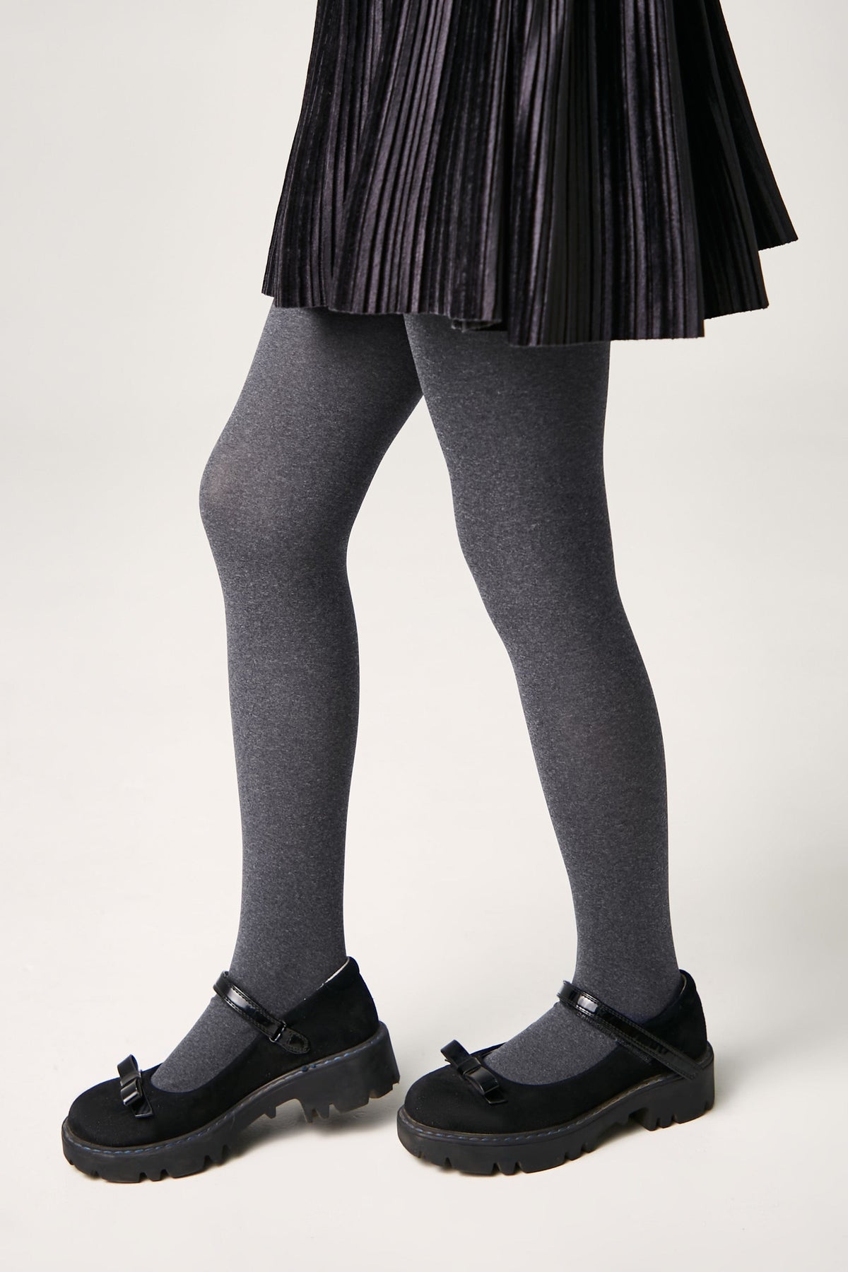 A person wears dark grey tights with black platform Mary Jane shoes featuring a bow detail and a pleated black skirt.