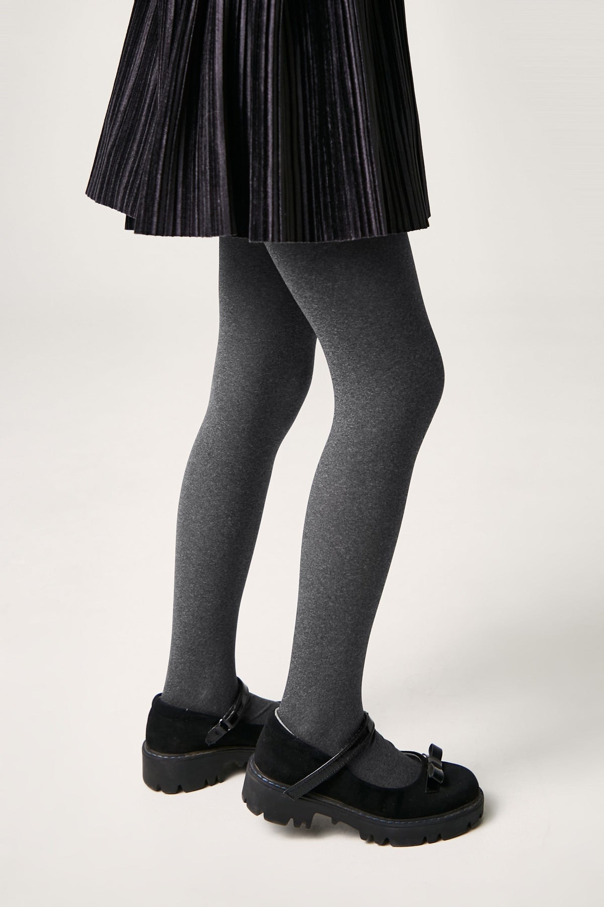 A person wearing a pleated black skirt, opaque charcoal grey tights, and black Mary Jane shoes with thick rubber soles.