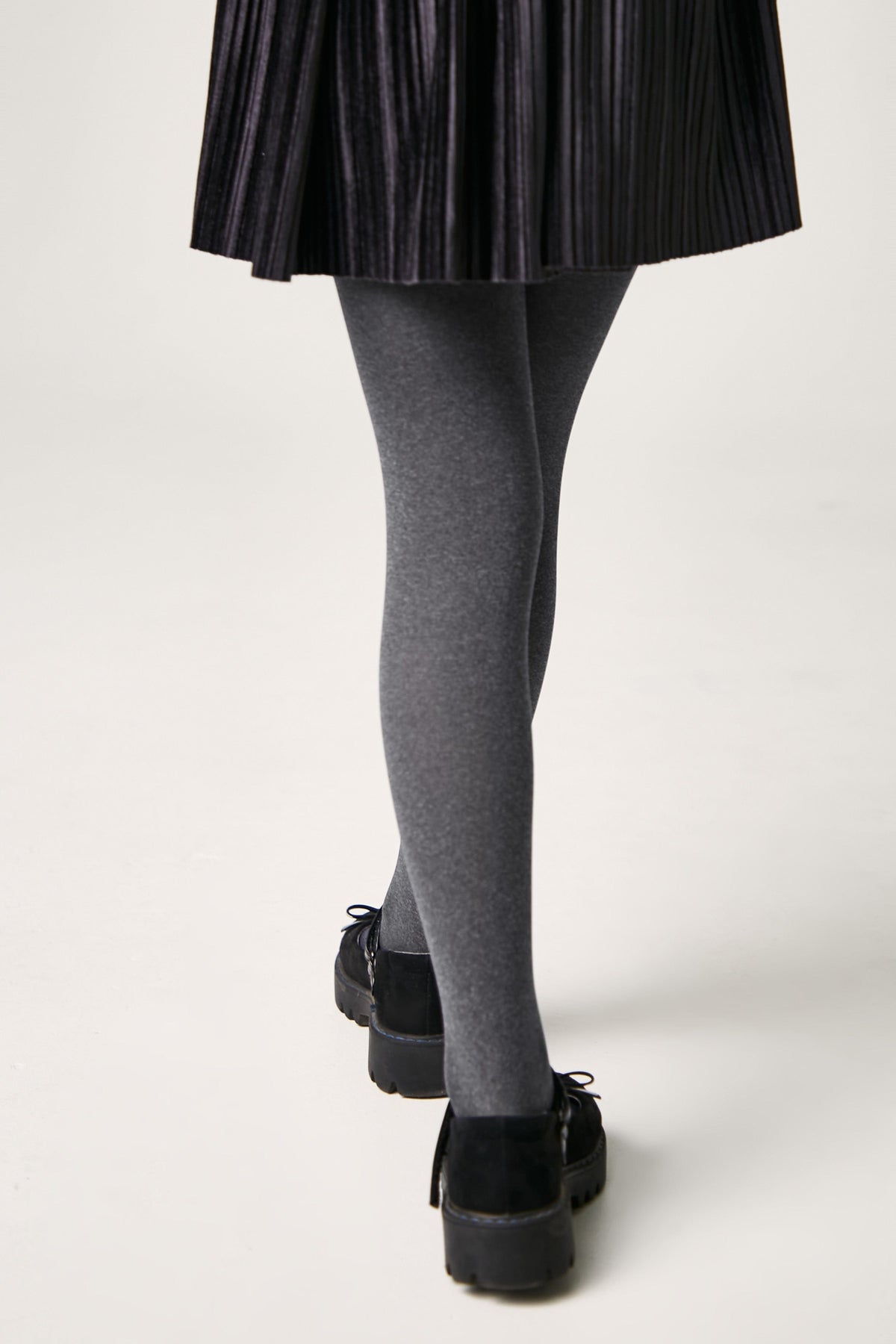 A person wears grey opaque tights with a black pleated skirt and black platform shoes against a plain white background.