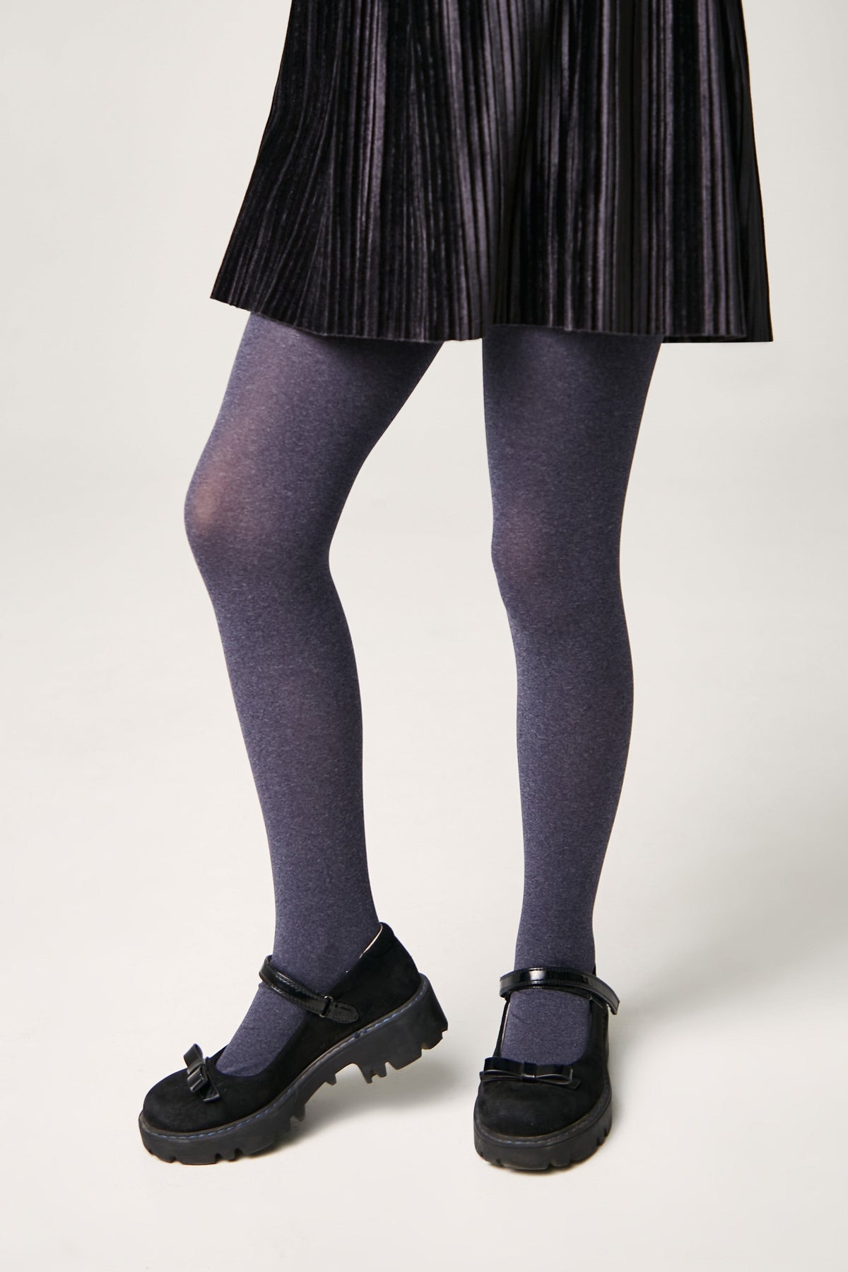 A person wears a black pleated skirt, grey tights, and black Mary Jane shoes with bow accents on a neutral background.