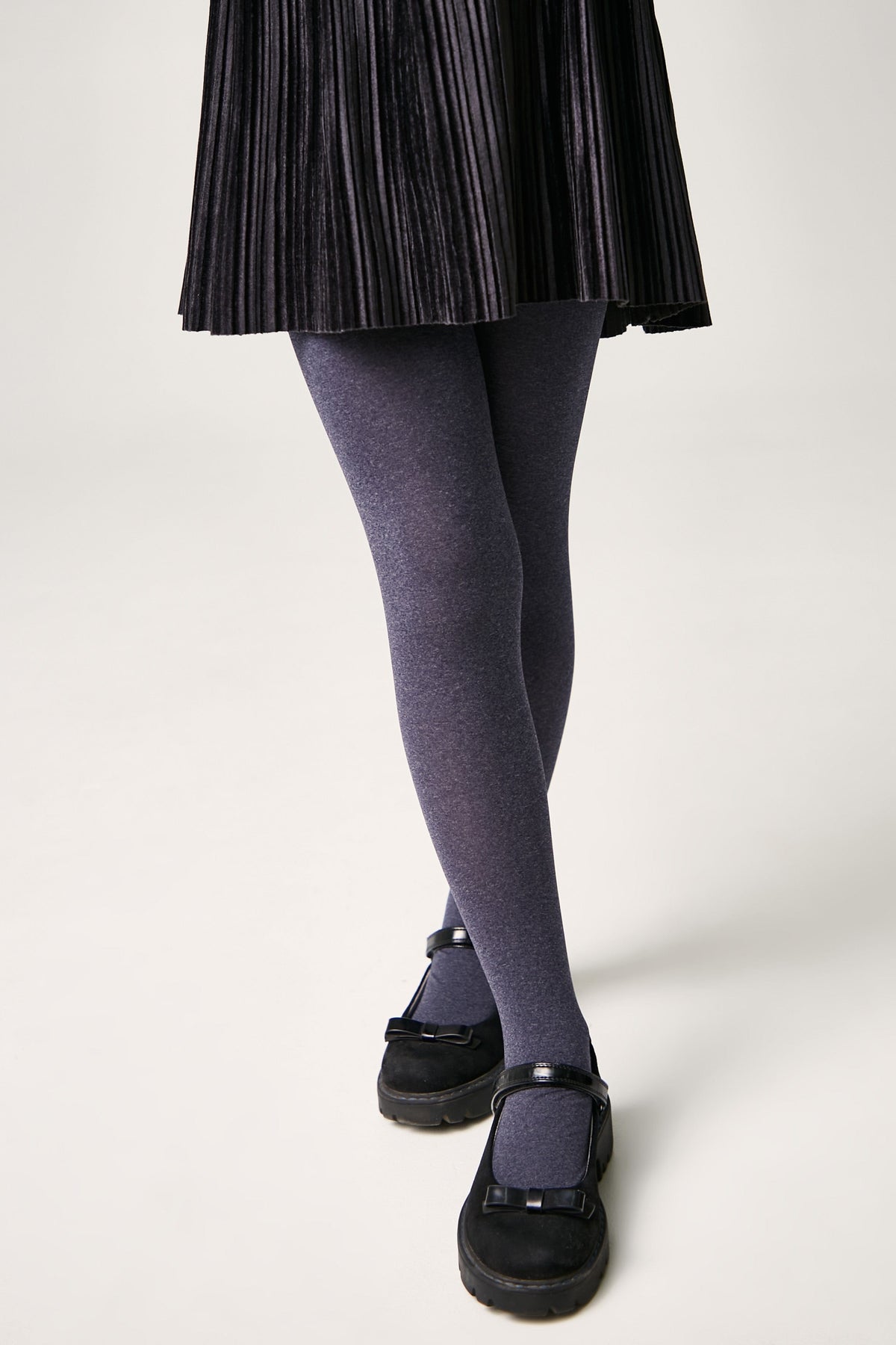 A pair of opaque purple-grey tights worn with a black pleated skirt and black mary jane shoes.