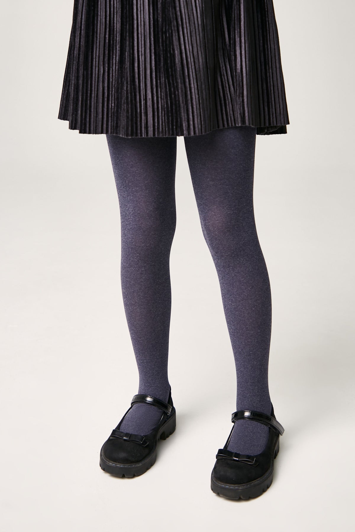 A person wears a pleated black skirt, heathered charcoal gray tights, and black patent leather mary jane shoes with a small bow detail.