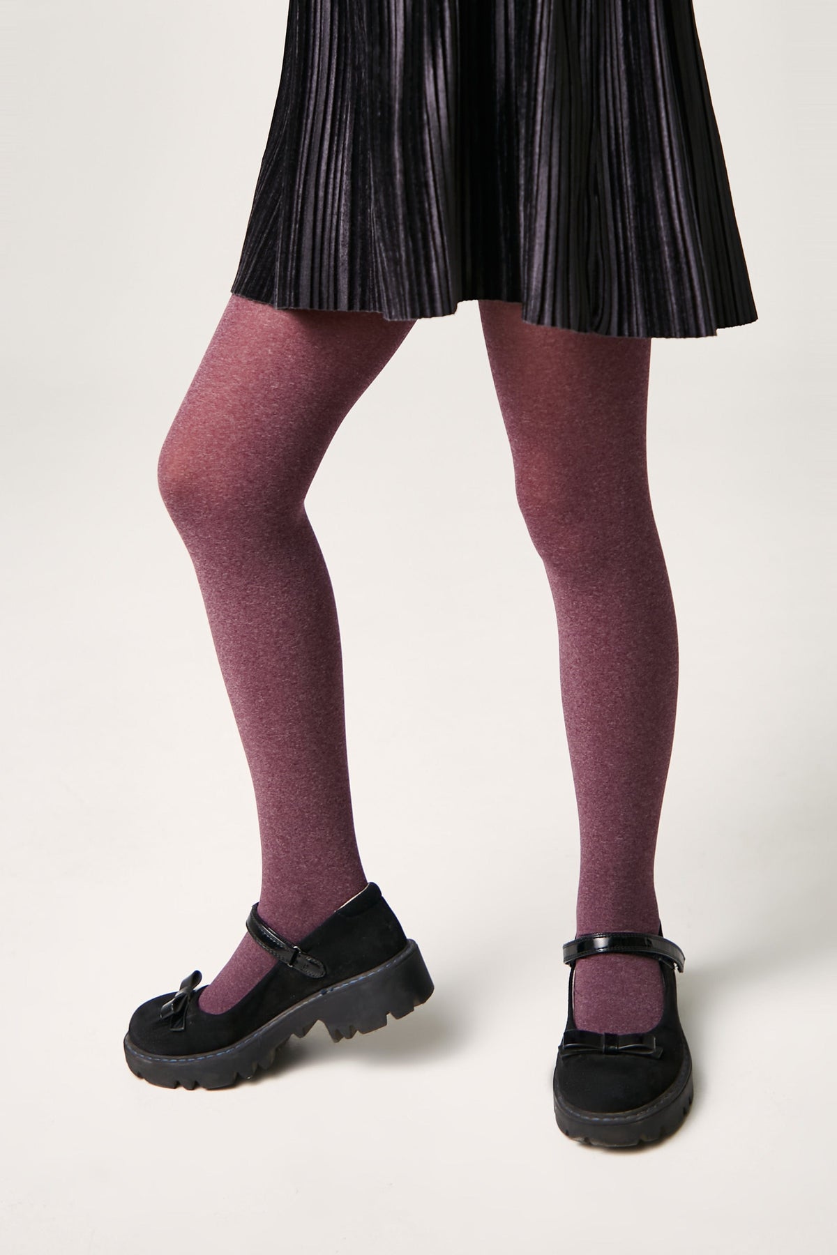 A person wears a black pleated skirt with opaque maroon tights and black Mary Jane shoes featuring a bow detail and chunky soles.
