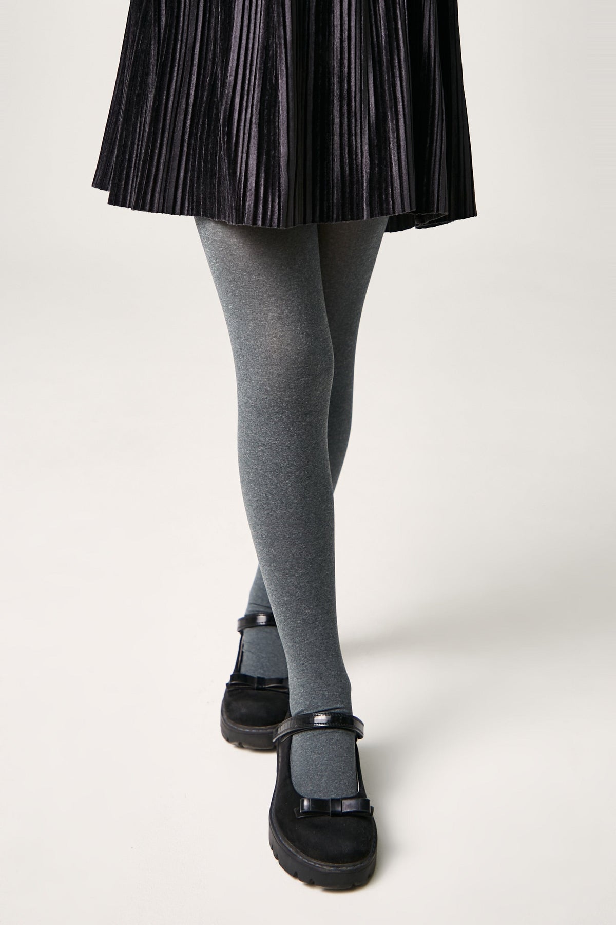 Grey tights worn with a black pleated skirt and black Mary Jane shoes against a white background.