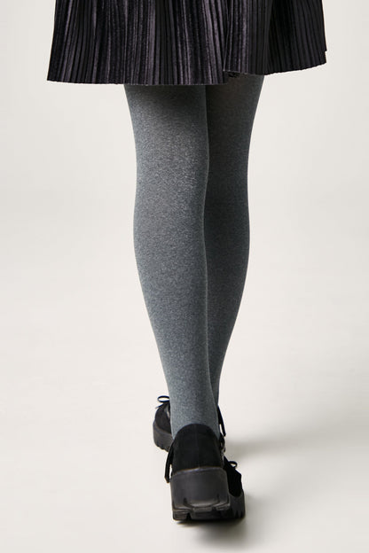 A person wears opaque grey tights paired with a black pleated skirt and black platform shoes, viewed from behind against a neutral background.