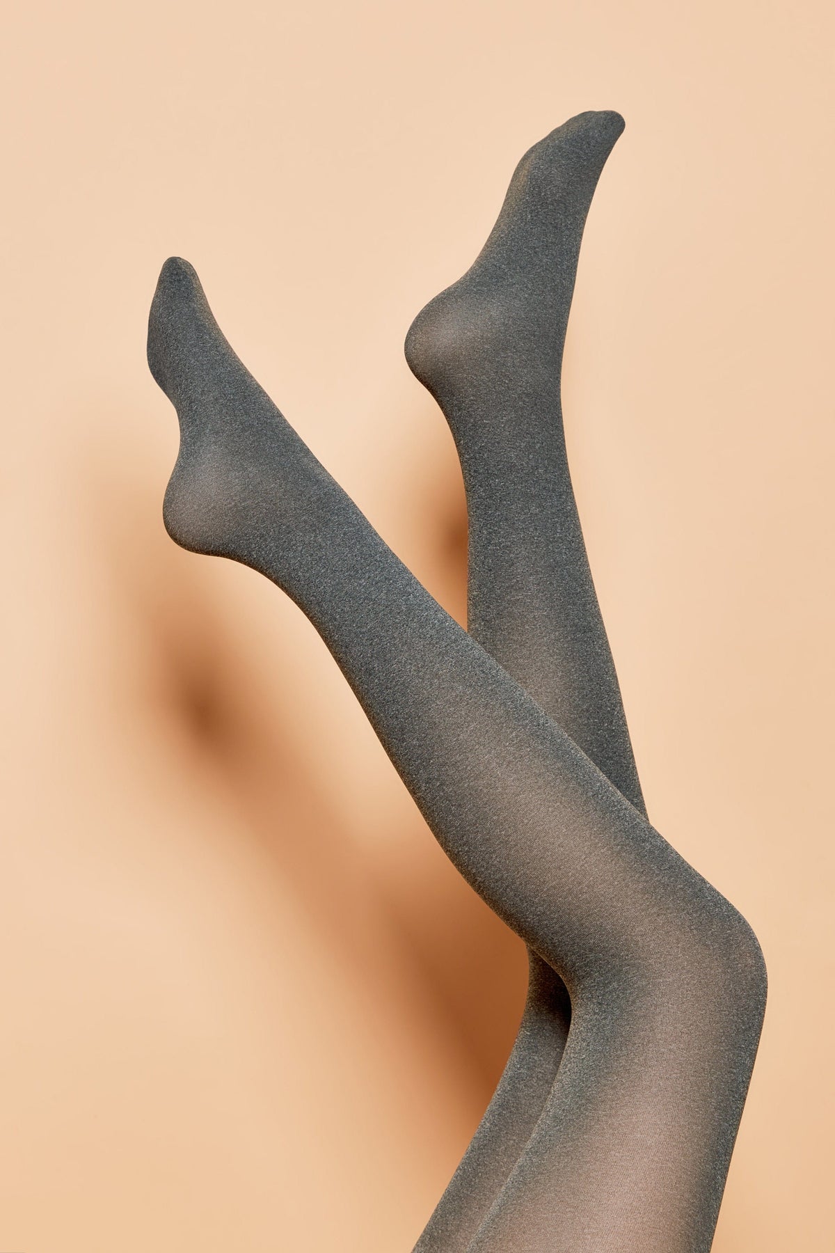 A pair of legs raised in the air, wearing charcoal grey opaque tights against a light peach-colored background.