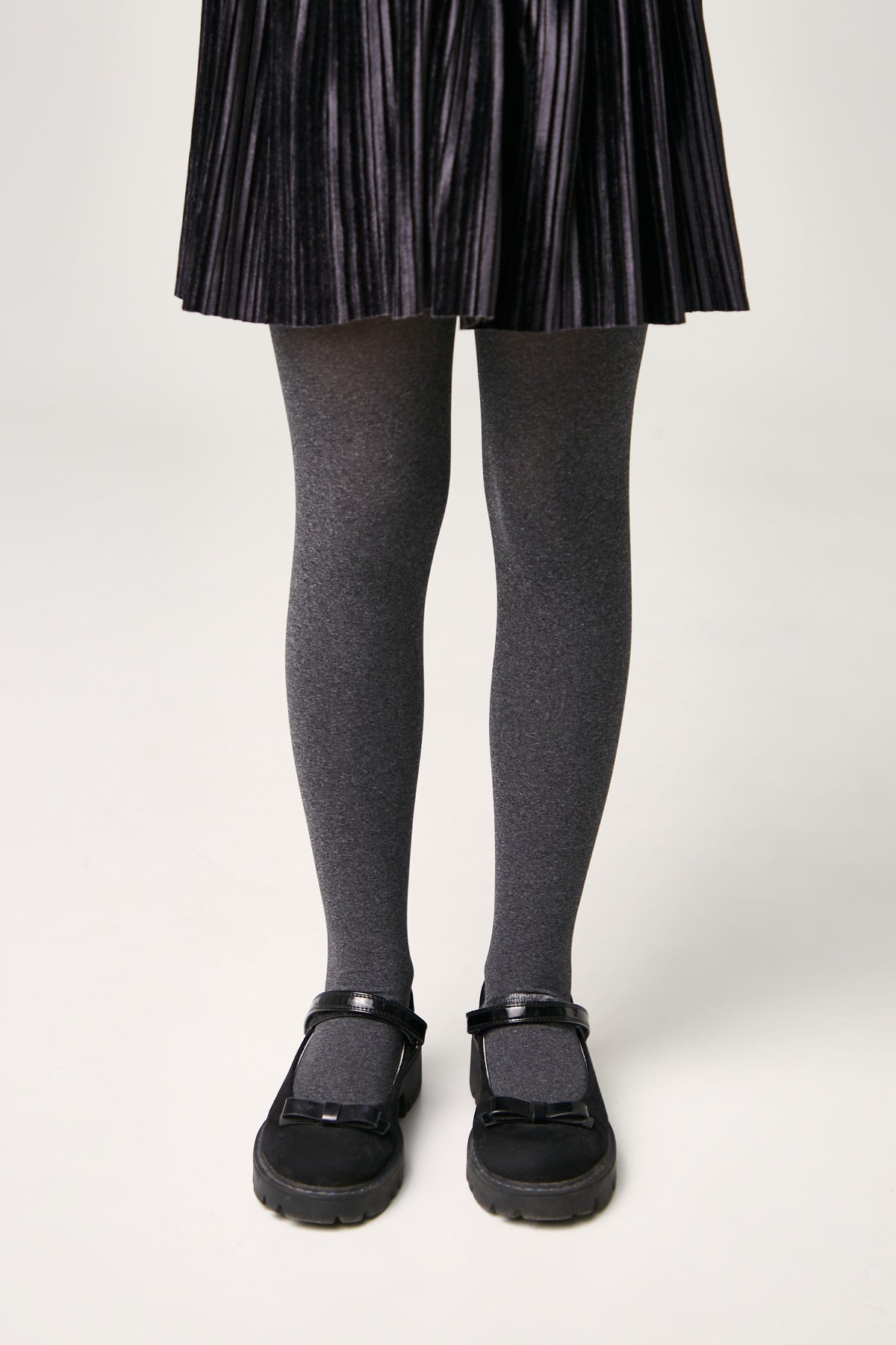 A pair of opaque grey tights worn with black Mary Jane style shoes featuring a bow detail on a plain white background.