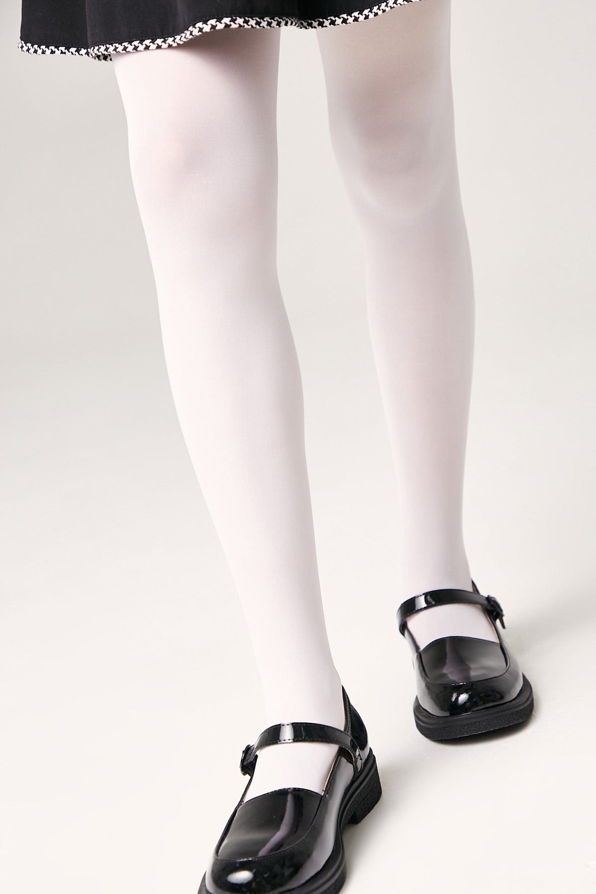 A pair of shiny black patent leather Mary Jane shoes worn with opaque white tights and a black skirt.