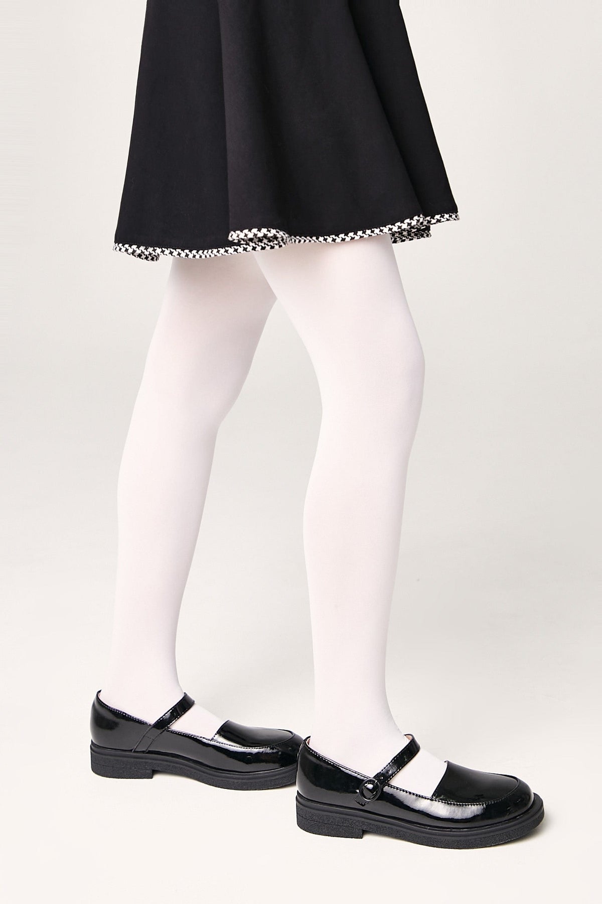 A person wearing a black flared skirt with a houndstooth-patterned hem, solid white tights, and black patent leather Mary Jane shoes against a plain off-white background.