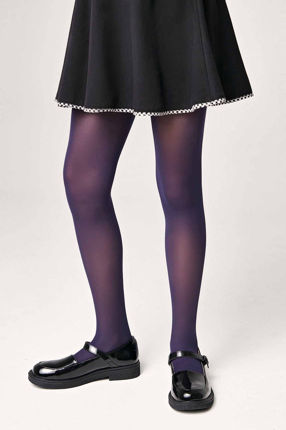 A person wears opaque dark purple tights paired with a black skirt featuring a decorative trim and black patent leather mary jane shoes.