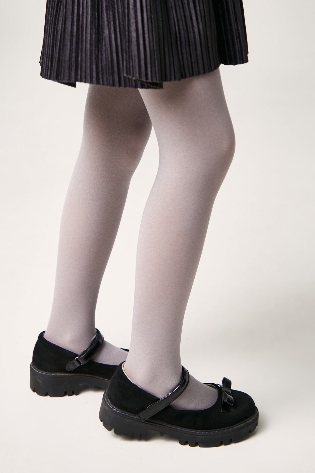 A close-up of a child's legs wearing grey tights and black flat shoes with a small bow detail, underneath a black pleated skirt against a white background.