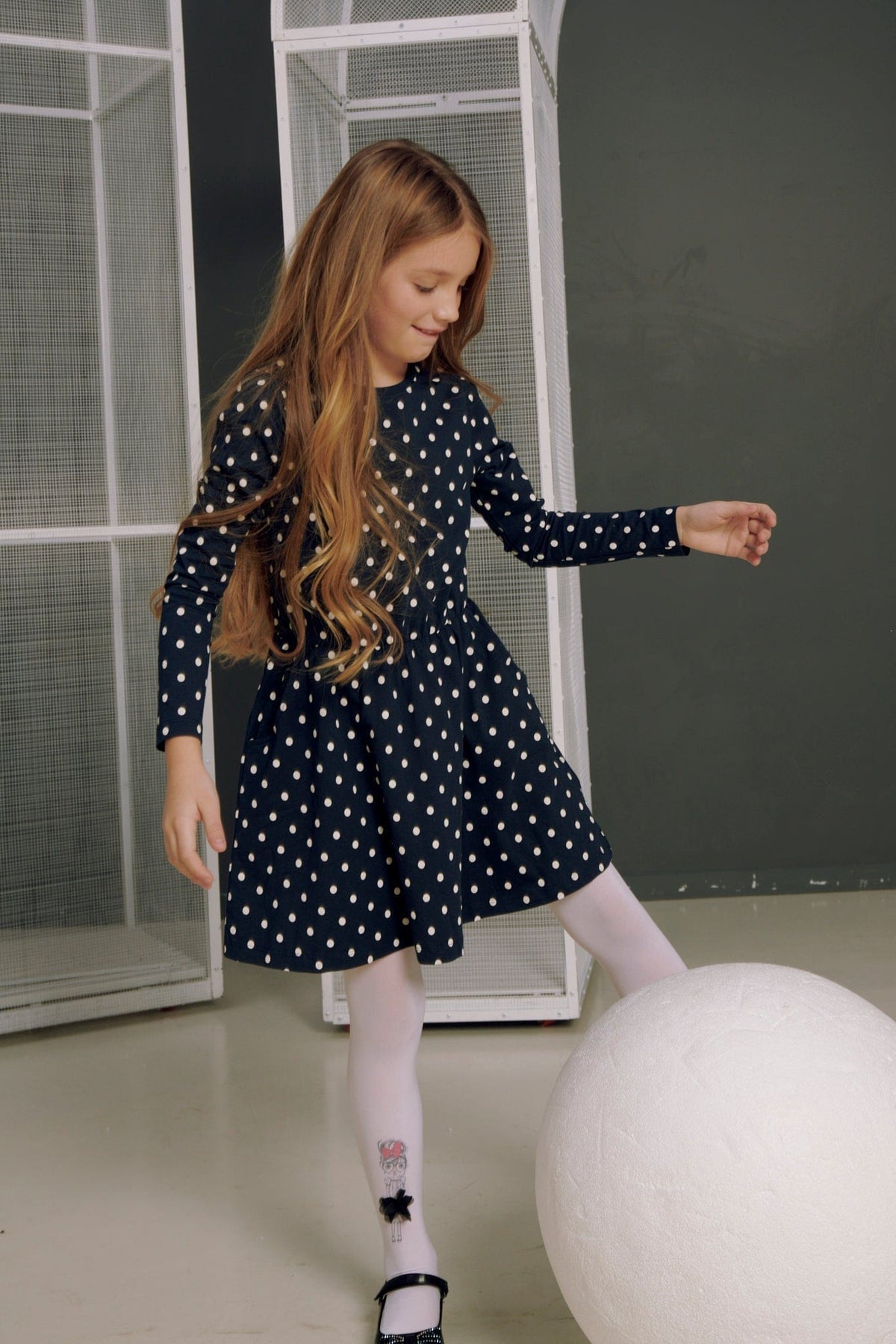 A young girl with long brown hair wears a long-sleeved navy blue dress with a white polka dot pattern and white tights while posing next to a large white sphere.