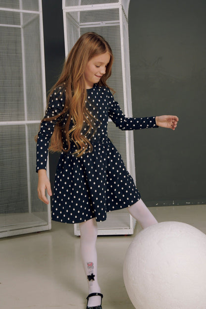 A young girl with long brown hair wears a long-sleeved navy blue dress with a white polka dot pattern and white tights while posing next to a large white sphere.