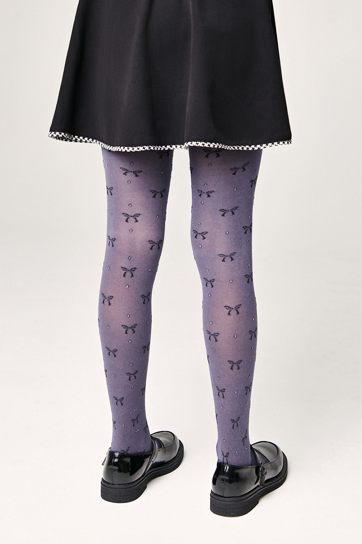 A person wears purple tights patterned with small dark bows and subtle sparkles, paired with a black skirt and black Mary Jane flats.